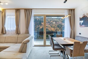 A modern living room with a sofa, dining table, chairs, and a view through a glass door to a forested area. - Apartment 'Elsa' with Mountain Views, Private Terrace and Wi-Fi (Fließ)