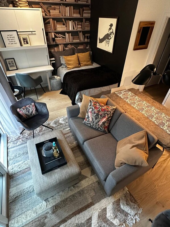 A modern living room with a grey sofa, a small coffee table, a bookshelf, a desk, and a bed with a black headboard. - Stunning studio with ocean view (Kristiansand)