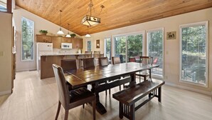 Uma ampla sala de jantar com mesa e bancos de madeira, uma cozinha com armários de madeira e janelas grandes que oferecem vista para o exterior.