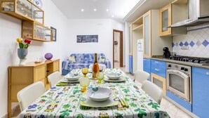 Ein Essbereich mit Tisch für vier Personen, eine Küche mit blauen Schränken und eine Couch im Hintergrund.