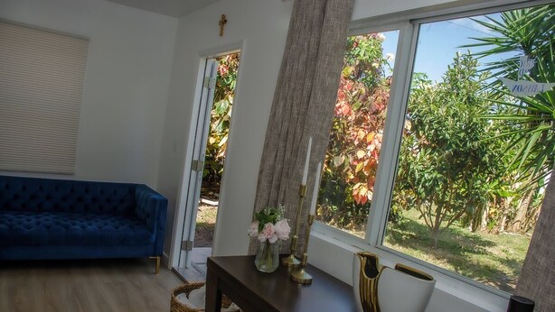 Een woonkamer met een blauwe bank, een donker houten tafeltje met een bloevas, en een uitzicht op een tuin door grote ramen.
