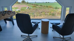 Ein modernes Zimmer mit einem großen achteckigen Fenster, zwei schwarzen Stühlen, einem Holztisch mit einer Tischfußballplatte und einem kleinen Hocker.