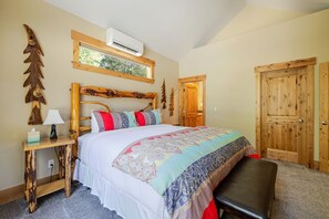 A bedroom with a wooden headboard, a bed with a patterned bedspread, a nightstand with a lamp, and a wooden door. - Aspen Place 3 Bedroom Home (Bend)