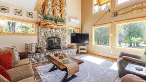 A cozy living room with a stone fireplace, large windows, and a television. - Aspen Place 3 Bedroom Home (Bend)