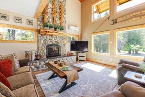 A cozy living room with a stone fireplace, large windows, and a television. - Aspen Place 3 Bedroom Home (Bend)