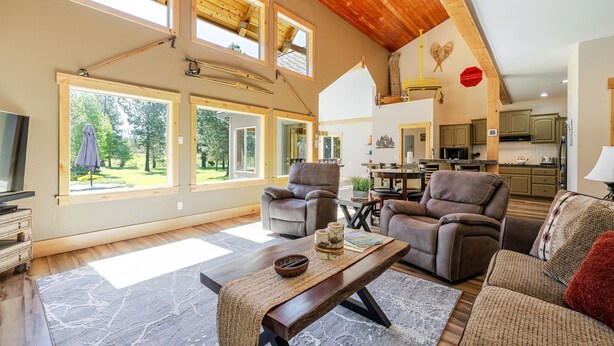 A spacious living area with a high ceiling, large windows, and a modern kitchen in the background. - Aspen Place 3 Bedroom Home (Bend)