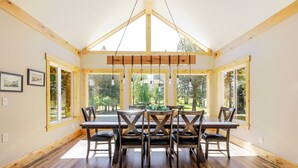 A dining area with a wooden table and chairs, large windows, and a high ceiling. - Aspen Place 3 Bedroom Home (Bend)