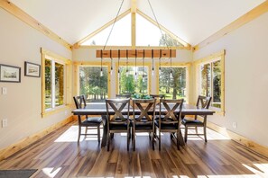 A dining area with a wooden table and chairs, large windows, and a high ceiling. - Aspen Place 3 Bedroom Home (Bend)