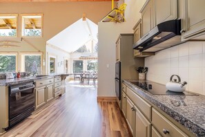 A spacious kitchen with wooden flooring, a built-in dishwasher, and a large window overlooking a dining area. - Aspen Place 3 Bedroom Home (Bend)
