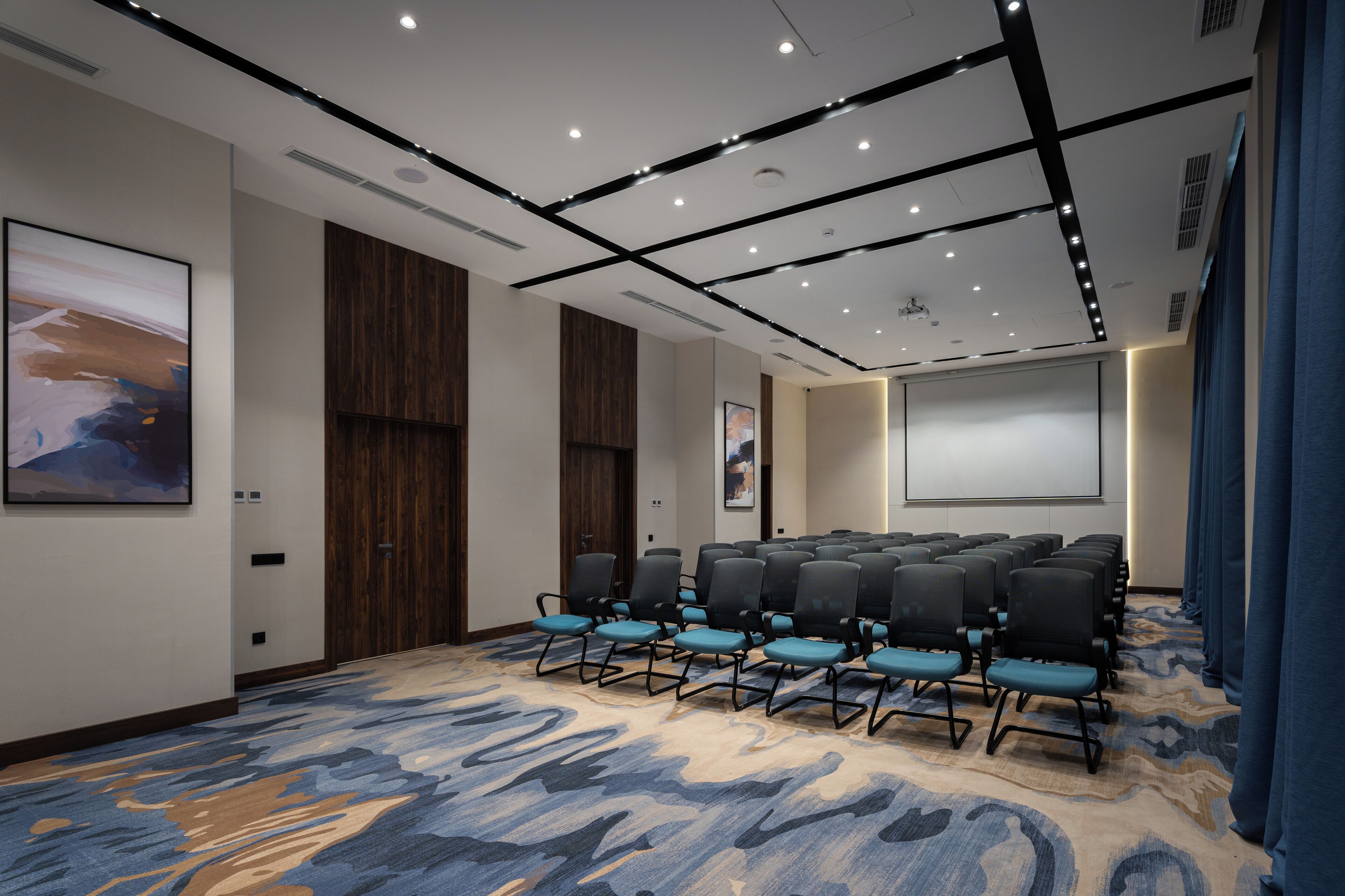 A conference room with rows of chairs, a projector screen, and a large abstract painting on the wall.