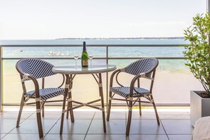 Een balkon met twee rieten stoelen en een tafel, met uitzicht op een strand met zeilboten.