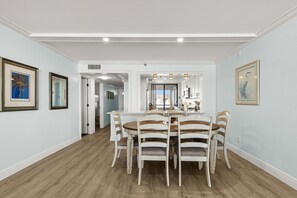 A dining area with a round table and six chairs, a kitchen in the background, and framed artwork on the walls. - ☀Beachfront Condo🌴Wrap Around Private Balcony🐬Redington Towers 3A🌞Heated Pool (Redington Shores)