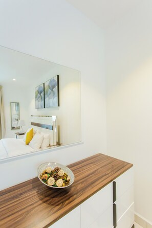 A modern hotel room with a wooden bedside table, a bowl of decorative items, a bed with white linens, and a large mirror reflecting the room. - Spacious One Bedroom at Embassy Gardens (Accra)
