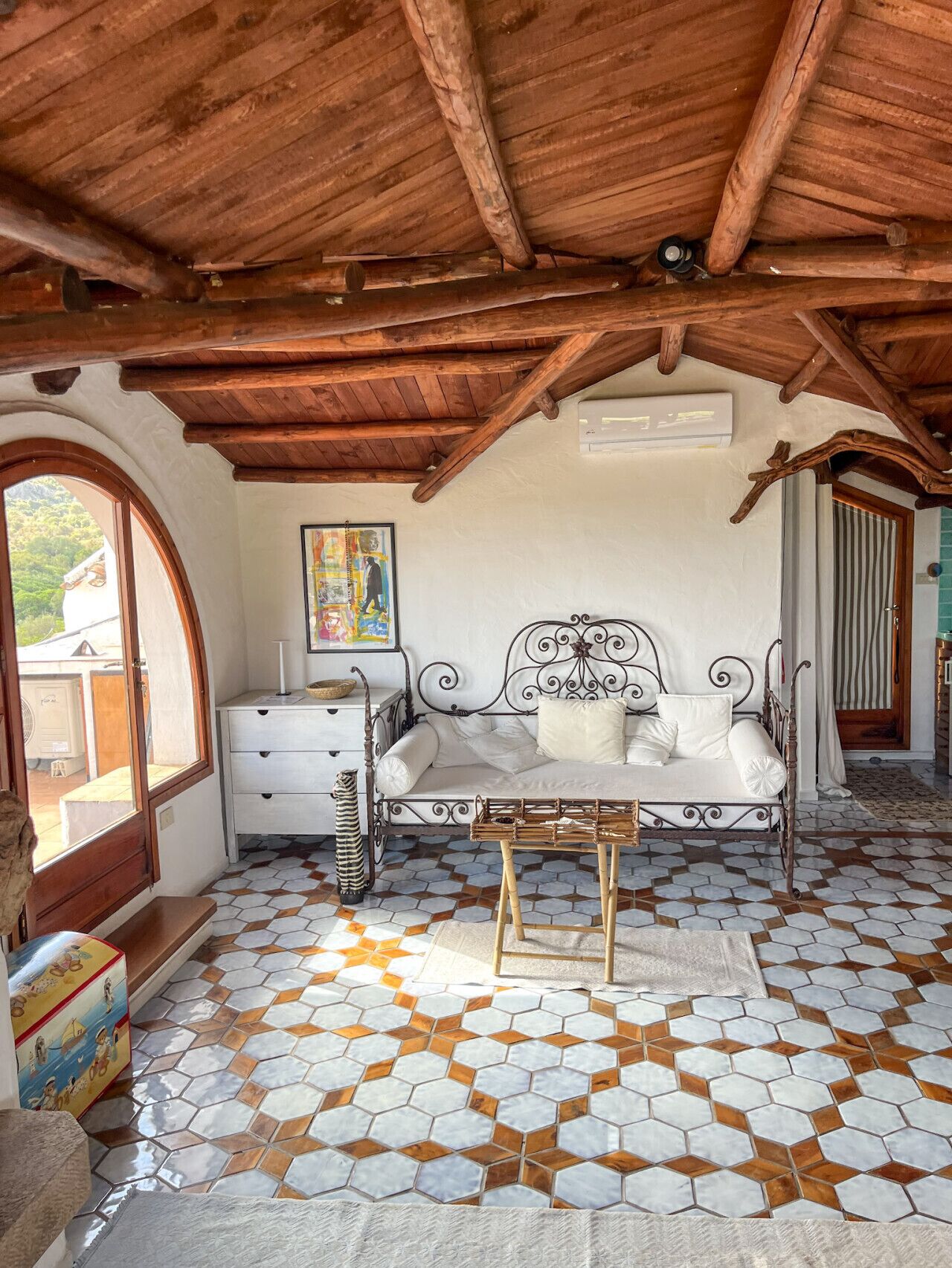 Une pièce chaleureuse avec un plafond en bois, un lit avec un cadre en métal, une table de chevet blanche et un tapis coloré.