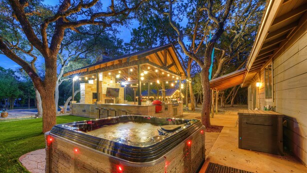 Un bain à remous doté d’une terrasse en bois et illuminé de guirlandes lumineuses, entouré d’arbres et d’un bâtiment avec un patio couvert.