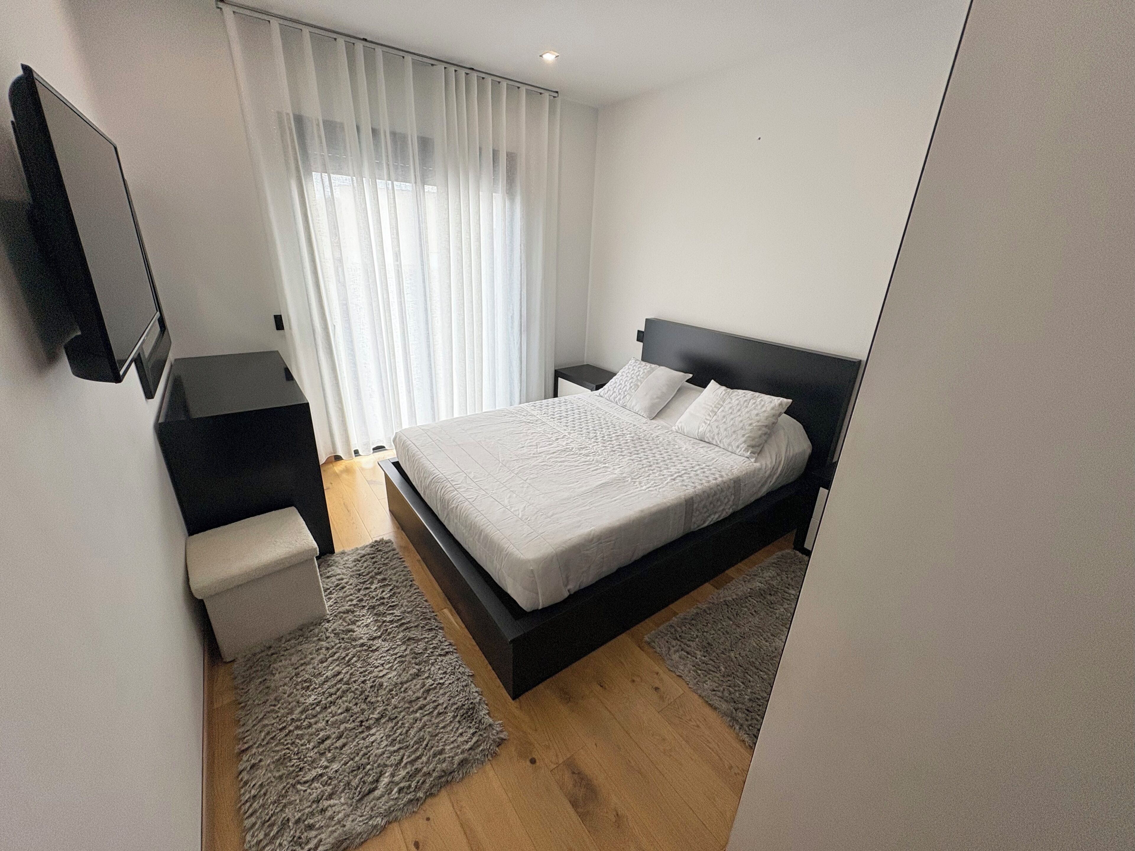 A modern bedroom with a bed, a TV mounted on the wall, and a window with sheer curtains.