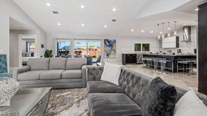 A modern living room with a grey sofa, a tufted armchair, and a dining area with a bar. - Heated Pool - Putting Green - 5 min From Beach! (Deerfield Beach)