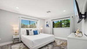 A modern bedroom with a large bed, bedside tables, a TV, and a view of greenery. - Heated Pool - Putting Green - 5 min From Beach! (Deerfield Beach)