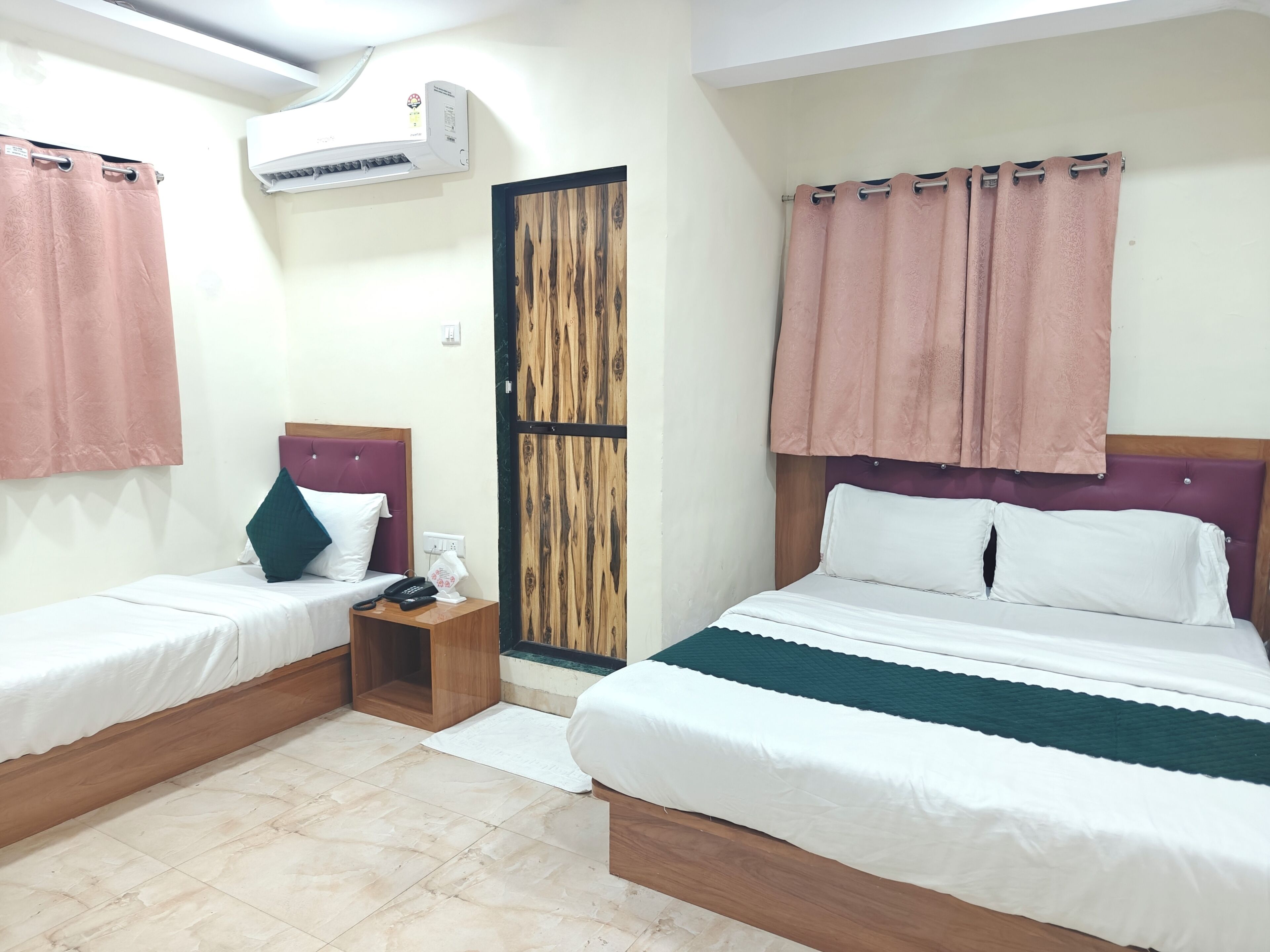 Habitación de hotel con dos camas, una puerta de madera, un equipo de aire acondicionado y una mesita de noche pequeña con un teléfono.