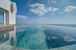 Una piscina infinita con agua azul cristalina que se extiende hasta el horizonte, un edificio moderno con un amplio ventanal y un cielo despejado.