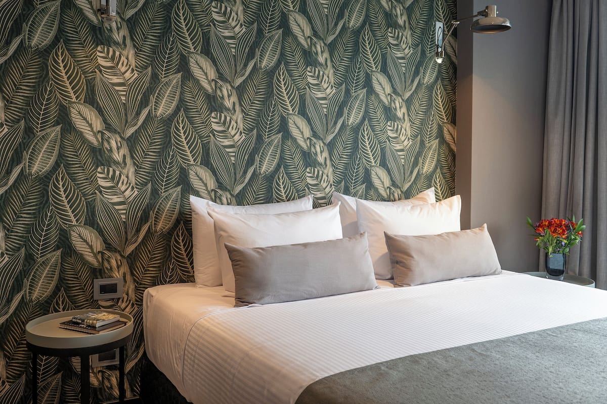 Een net opgemaakt bed met witte lakens en meerdere kussens, een klein rond tafeltje met een boek en een afstandsbediening, en een wand met een bladerpatroon.