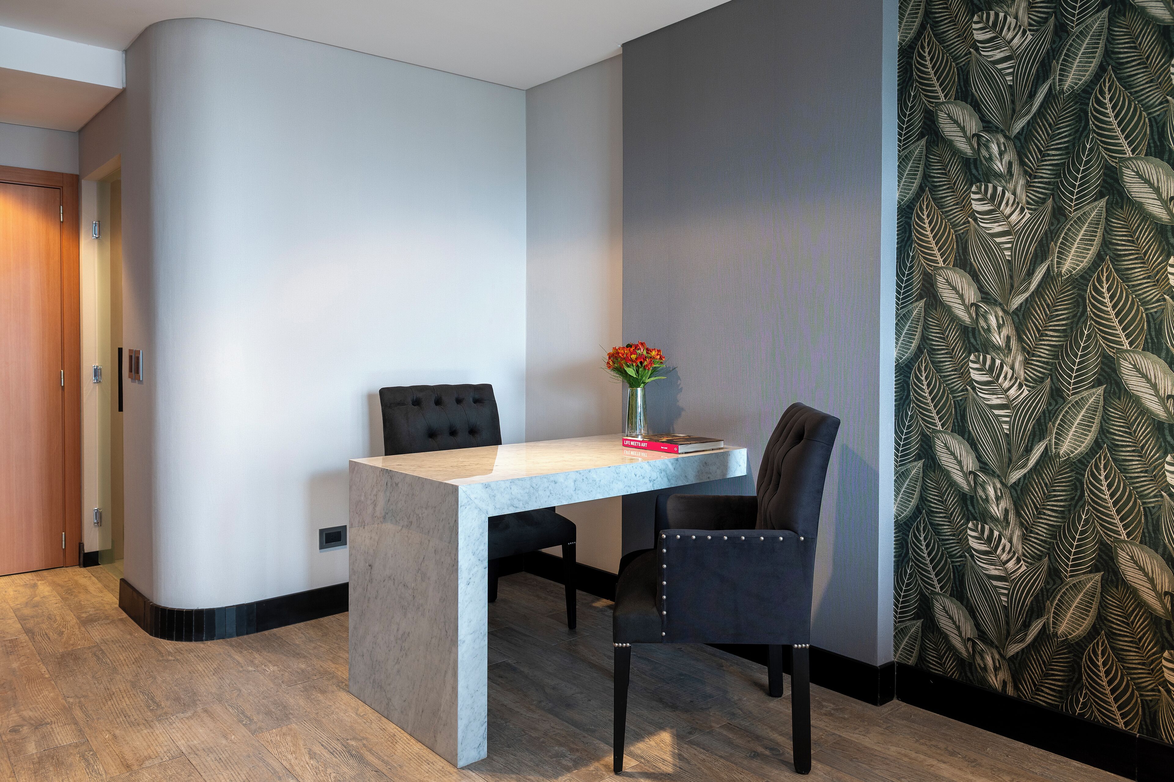 A modern room with a marble table, two black upholstered chairs, a vase with flowers, and a leaf-patterned wallpaper.
