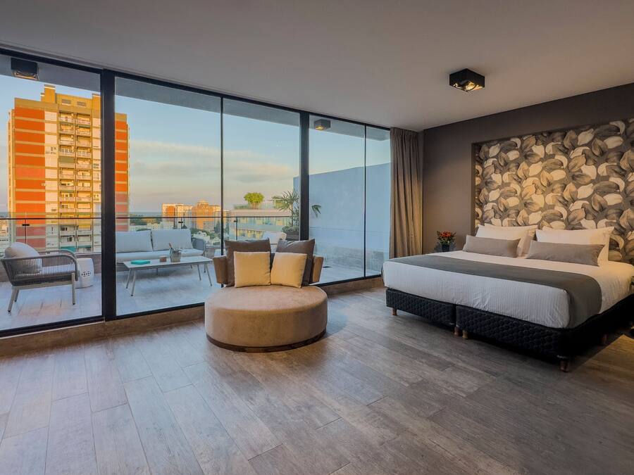 A modern bedroom with a large bed, a round ottoman, and a sofa. It features a large window with a city view.