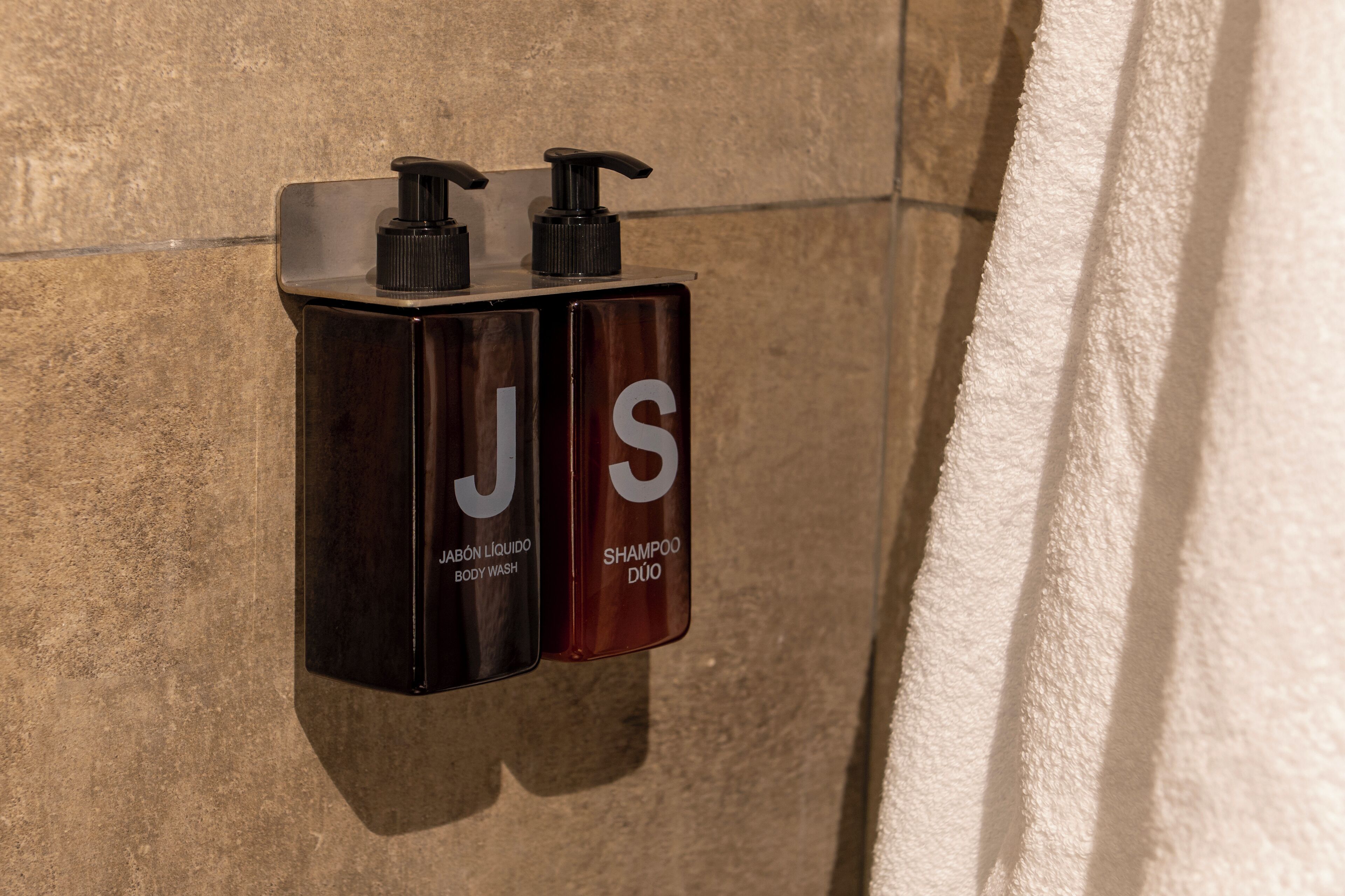 Deux bouteilles noires équipées de pompes, portant les étiquettes « JABÓN LÍQUIDO BODY WASH » et « SHAMPOO DUO », sont fixées au mur.