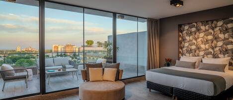 A modern bedroom with a large bed, a round ottoman, and a sofa. It features a large glass window with a city view.