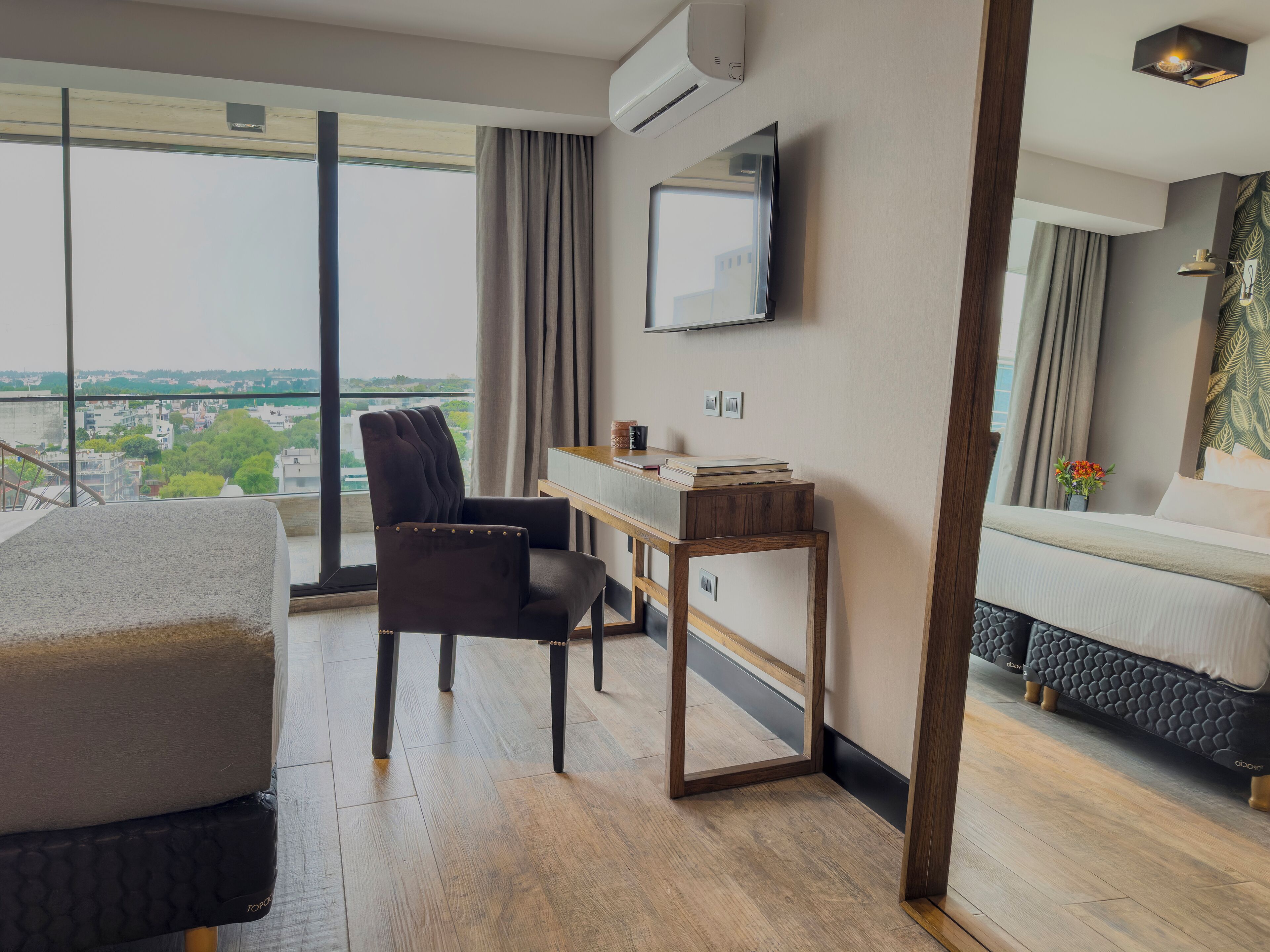 A modern hotel room with a large window, a flat-screen TV, a desk, a comfortable chair, and a bed.
