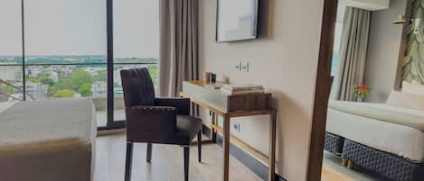 A modern hotel room with a large window, a flat-screen TV, a desk, a comfortable chair, and a bed.