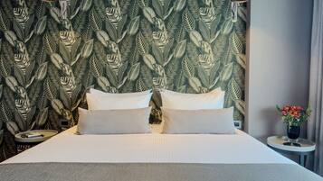A neatly made bed with white linens and pillows, a green leaf-patterned wallpaper, and a small table with a vase of flowers.