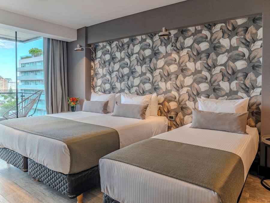 Una camera d'albergo con due letti, un'ampia finestra vista edifici, una parete decorata e un tavolino con un telefono.