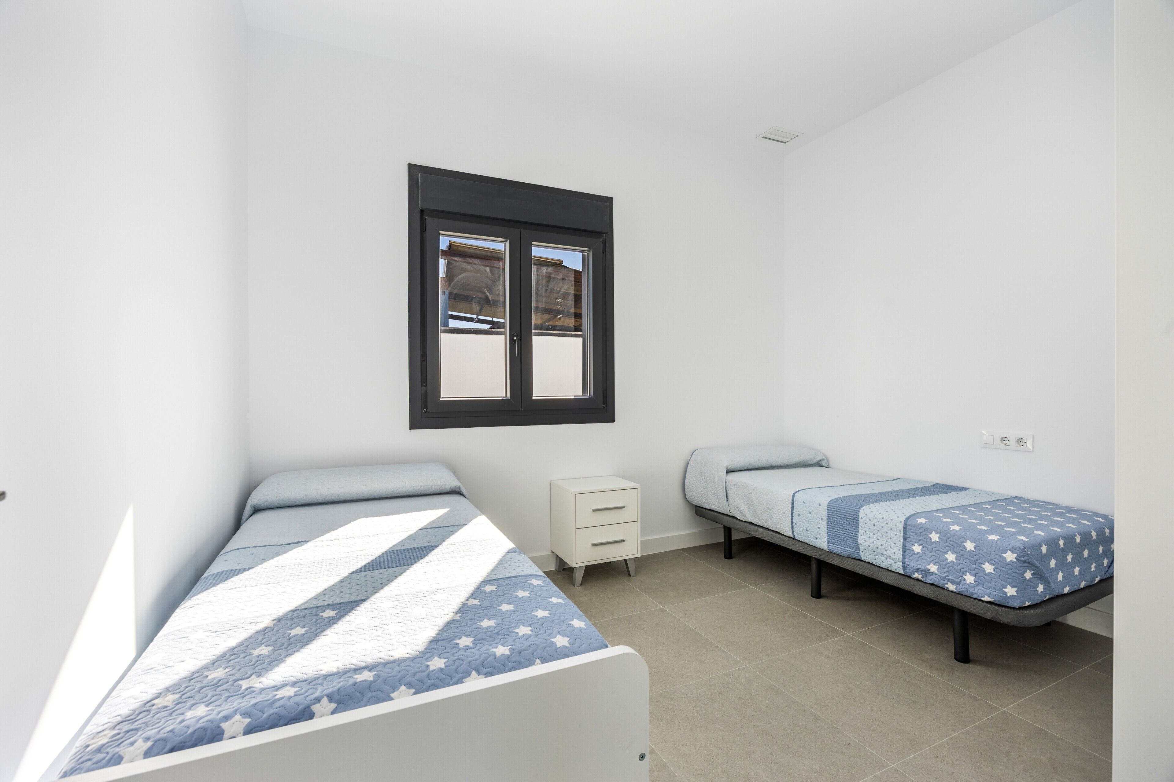 Une chambre avec deux lits simples, une petite table de chevet, une fenêtre et une prise électrique au mur.