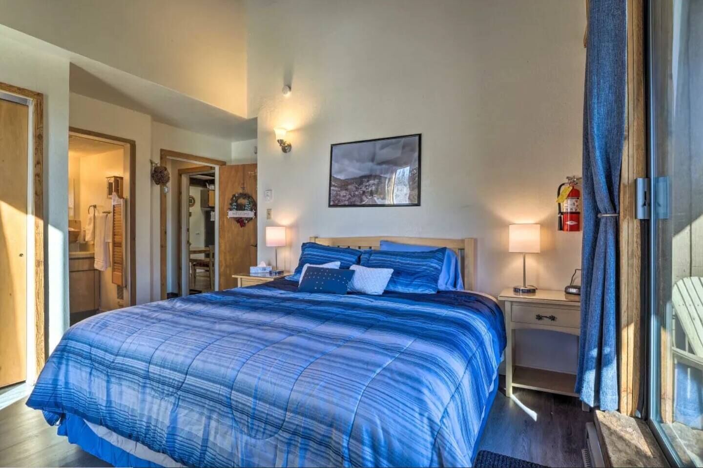 A bedroom with a large bed, bedside lamps, a framed picture on the wall, and a view of the outside.