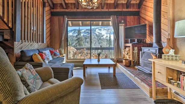 A cozy cabin interior with a sofa, a table, a fireplace, and a large window offering a mountain view. - Aspen View Lodge 4 bed/3 ba, Sauna, W/D, Views (Brian Head)