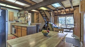 A rustic interior with a wooden dining table, a kitchen area, and a staircase leading to a balcony with a view of trees. - Aspen View Lodge 4 bed/3 ba, Sauna, W/D, Views (Brian Head)
