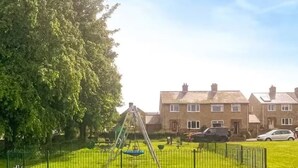 Ein Spielplatz mit Schaukel und Rutsche, umgeben von einem eingezäunten Rasenbereich und Wohnhäusern im Hintergrund.