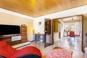 Uma sala de estar moderna com televisão de tela plana, móveis de madeira, um sofá vermelho, um cobreleira floral e vista para uma área de jantar com mesa e cadeiras.