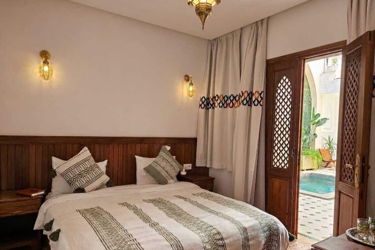 A hotel room with a bed, wooden headboard, two wall sconces, a patterned bedspread, a wooden nightstand, and a door leading to an outdoor area.