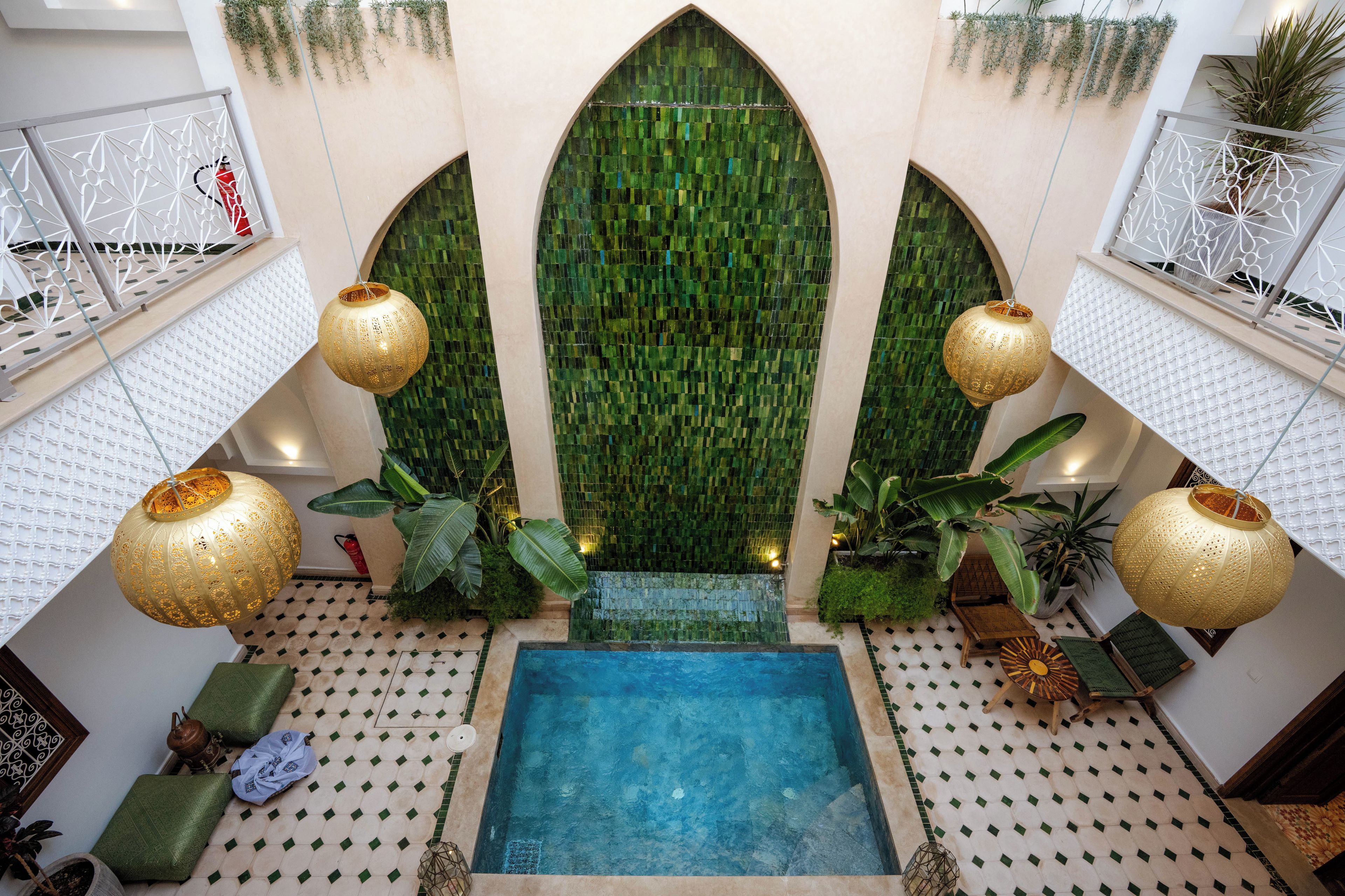 Un área de piscina con una piscina rectangular central, rodeada de suelo estampado y flanqueada por paredes de mosaico verde y faroles colgantes.