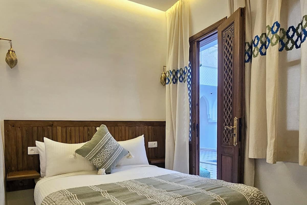 A bedroom with a wooden headboard, a large bed with patterned bedding, a bench, and a view of the outside through a door.