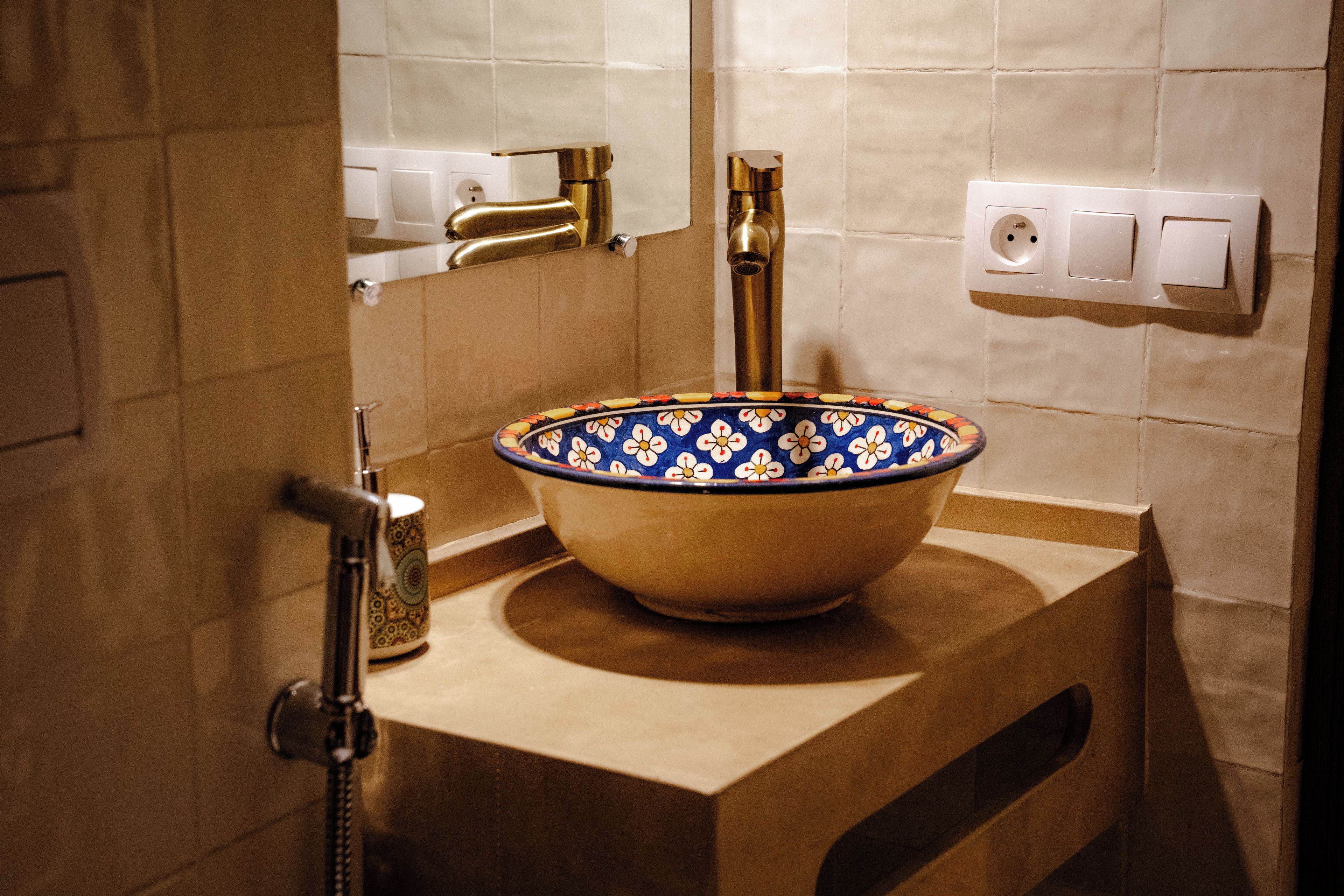 Un baño con encimera color beige, un lavamanos tipo cuenco con estampado azul y blanco, grifo dorado y ducha de mano.