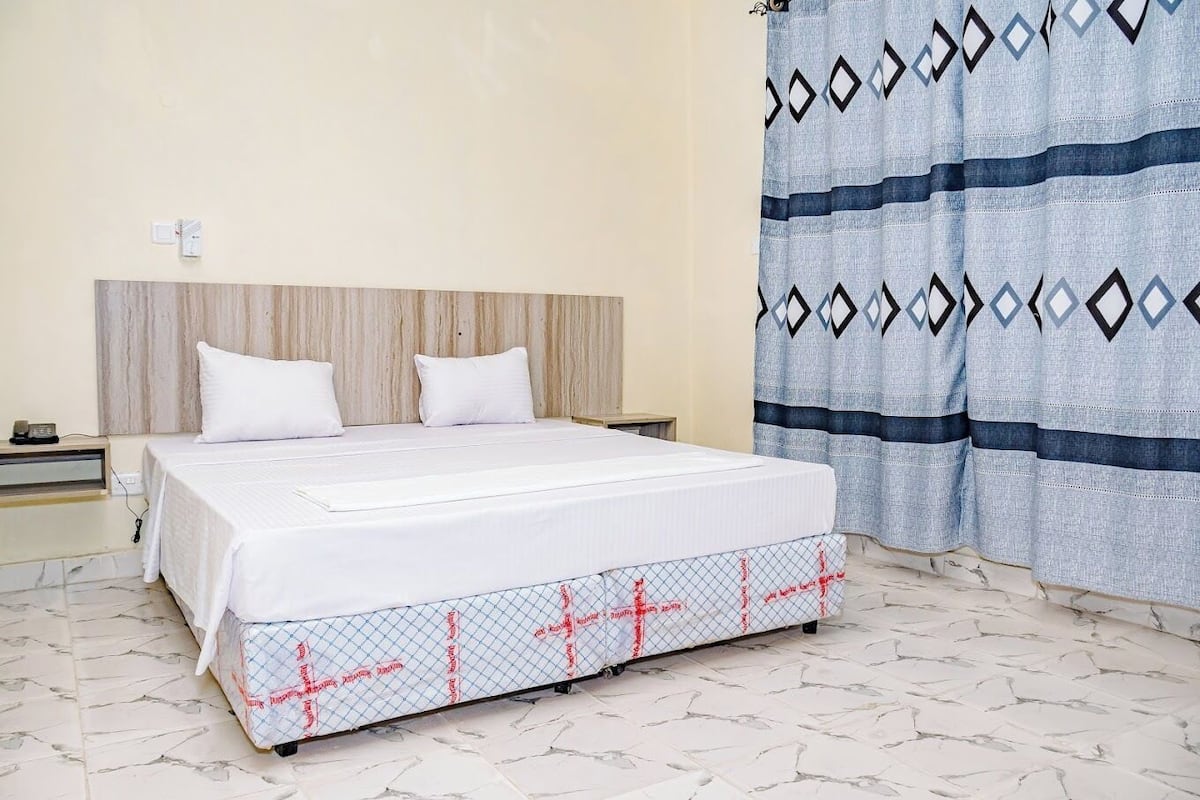 A bedroom with a bed, two white pillows, a wooden headboard, a nightstand, a power outlet, and a patterned curtain.