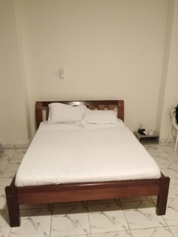 A single bed with a wooden frame and white bedding in a room with white walls and tiled flooring.