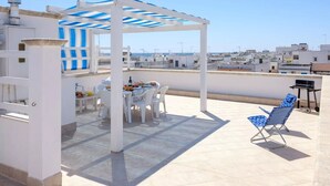 Una terraza en la azotea con zona de estar cubierta, una mesa con sillas y una parrilla.
