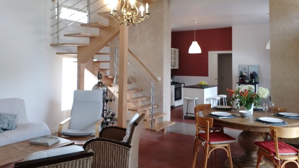 Un vaste espace de vie doté d’un haut plafond, de poutres en bois, d’un escalier, d’une table à manger pour quatre personnes et d’une cuisine à l’arrière-plan.