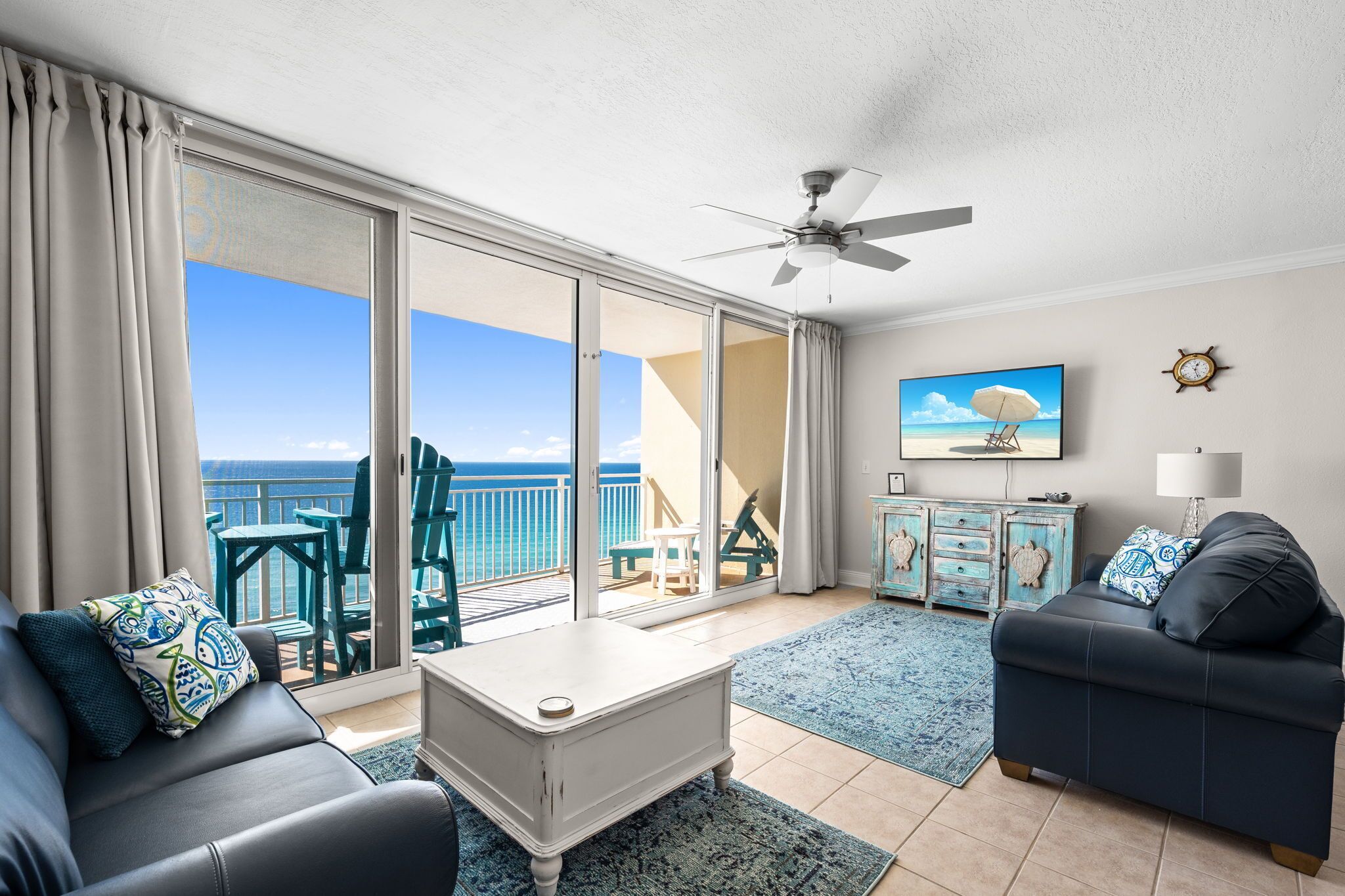 A living room with a view of the ocean, a ceiling fan, a TV, and a clock on the wall.