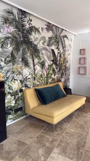 A yellow sofa with a dark cushion in a room with a tropical-themed mural on the wall.