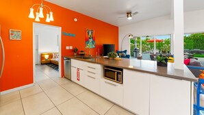 A modern kitchen with white cabinets, an orange accent wall, a built-in microwave, and a dining area with a view of the outdoors. - 30+ Stay! Perfect For Families, W/Pool & Hot Tub (Palm Springs)